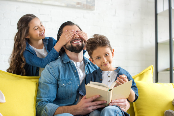 милая маленькая дочь закрывает глаза на счастливого отца, в то время как отец и сын читают книгу на диване дома
 - Фото, изображение