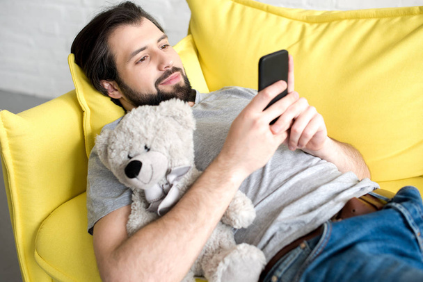 усміхнений молодий чоловік використовує смартфон, лежачи на дивані і обіймаючи плюшевого ведмедя
 - Фото, зображення