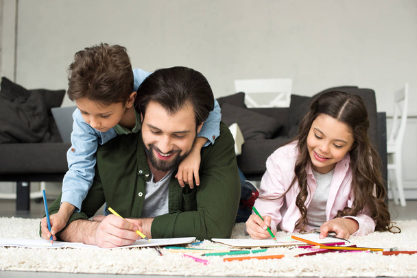 glücklicher Vater mit niedlichen Kindern, die auf Teppich liegen und mit Buntstiften zeichnen  - Foto, Bild