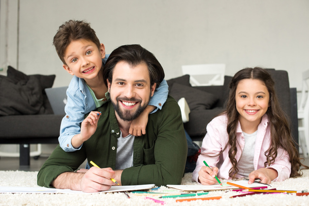 счастливый отец с милыми маленькими детьми, улыбающимися в камеру во время рисования цветными карандашами дома
 - Фото, изображение
