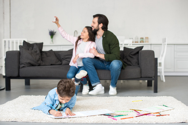 happy father and daughter sitting on sofa and taking selfie with smartphone while little boy drawing on carpet at home - 写真・画像