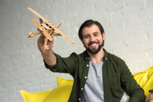 close-up view of happy young man playing with wooden toy plane and smiling at camera - Fotografie, Obrázek