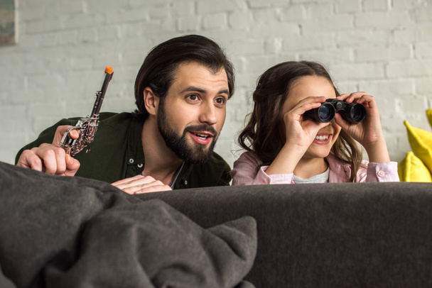lachende vader en dochter met verrekijkers en speelgoed pistool verbergen achter Bank en thuis samenspelen - Foto, afbeelding