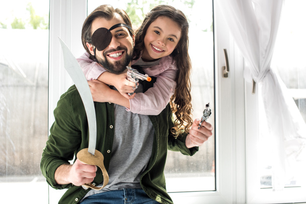 glücklicher Vater und Tochter lächeln beim gemeinsamen Spielen in die Kamera und geben vor, zu Hause Piraten zu sein  - Foto, Bild