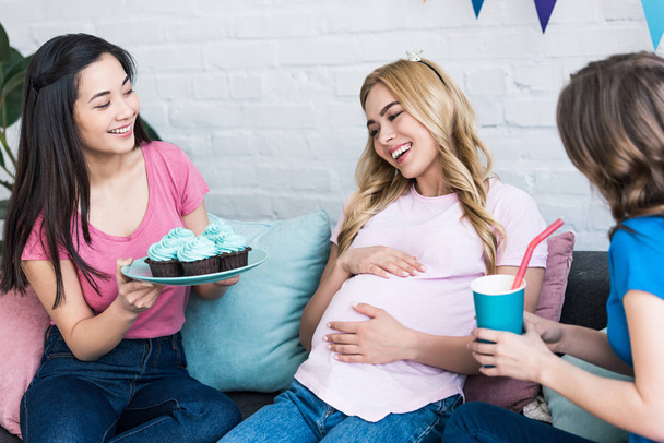 multicultural women proposing pregnant friend cupcakes and drink during baby-party - Foto, Imagen