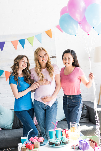 amigos multiculturales sonrientes y mujer embarazada de pie con globos en la fiesta del bebé
 - Foto, Imagen