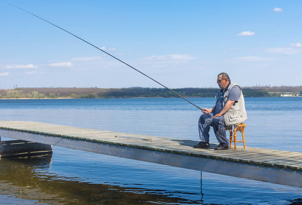 Samotny starszy rybak siedzi na stołek wiklinowe na molo z prętem i gotowe do połowu fis - Zdjęcie, obraz