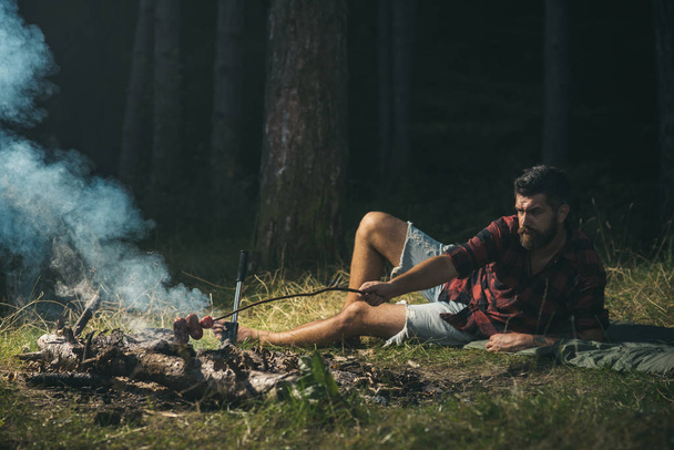 Lone hiker wandering in woods, camping concept.. Hipster cooking sausages over fire. Bearded barefoot man lying on grass. - Фото, зображення