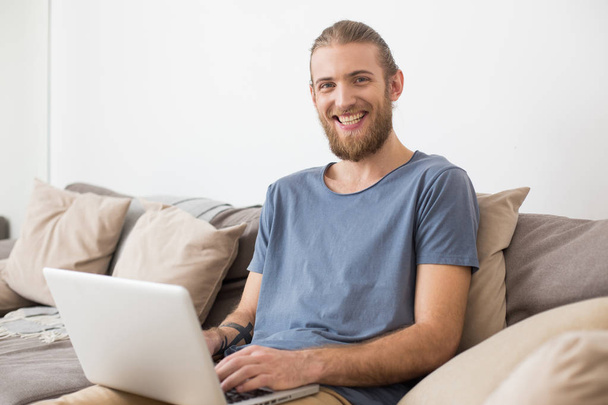 Retrato de un joven sonriente sentado en un gran sofá gris con portátil y mirando alegremente en cámara aislada
 - Foto, Imagen