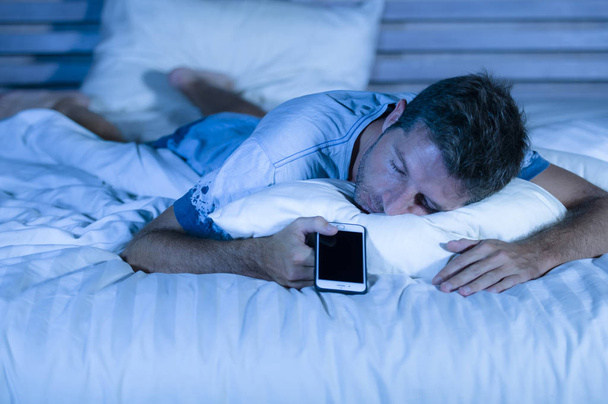 aantrekkelijke moe man in bed in slaap vallen tijdens het gebruik van de mobiele telefoon nog steeds houdt de cellulaire in zijn hand tijdens het slapen in internet en sociale media-verslaving - Foto, afbeelding