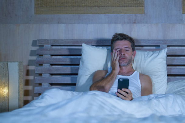 portrait of young attractive man lying in bed tired and relaxed using mobile phone looking sleepy at his apartment bedroom in internet addiction - Foto, afbeelding