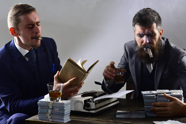 Company engaged in illegal business. Men sitting at table with piles of money and typewriter. Illegal business concept. Businessmen discussing illegal deal while drinking and smoking, grey background. - 写真・画像