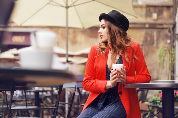 Young stylish beautiful woman sitting in city cafe in red jacket, street style, drinking aromatic coffee. Elegant girl in hat smile. Sweet breakfast, happy face, outdoor hipster portrait, fashion girl - Photo, Image