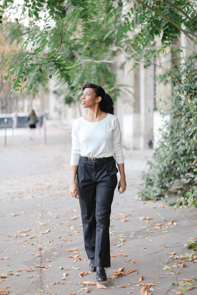 Mujer afroamericana encantadora caminando en la ciudad cerca de árboles verdes y el edificio, con blusa blanca
. - Foto, imagen