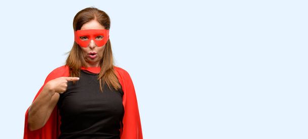 Μέση ηλικία σούπερ ήρωας γυναίκα που φοράει κόκκινη μάσκα και Ακρωτήριο ευχαριστημένοι και έκπληκτοι επευφημίες εκφράζοντας wow χειρονομία, δείχνοντας με το δάχτυλο απομονωμένο γαλάζιο φόντο - Φωτογραφία, εικόνα