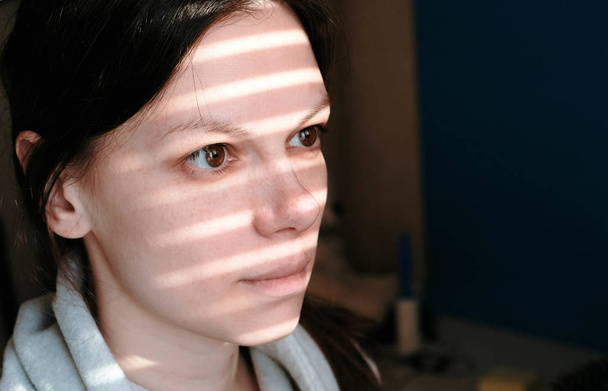 Close-up of a brunette womans face with stripes of light from the window on her face. - Photo, Image