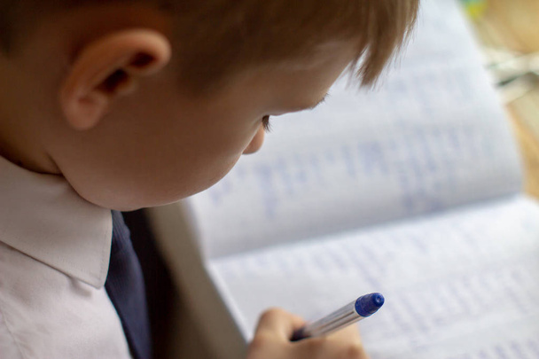 家庭教育。放課後の家の仕事。伝統的な白いメモ帳の紙に手で英単語を書くペンを持つ少年.  - 写真・画像