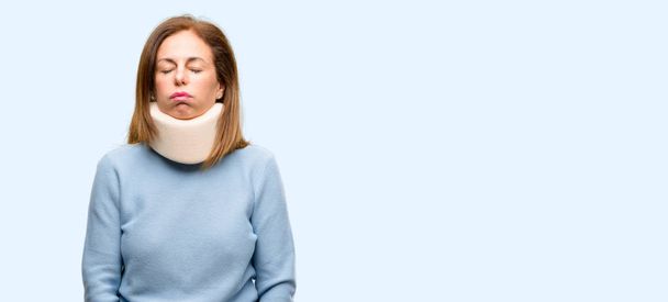 Τραυματισμένη γυναίκα φοράει κολάρο στο λαιμό τιράντες με υπνηλία έκφραση, είναι καταπονημένος και κουρασμένος απομονωμένες μπλε φόντο - Φωτογραφία, εικόνα