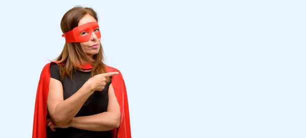 Супергероиня средних лет, женщина в красной маске и накидке, указывающая на сторону пальца на голубом фоне
 - Фото, изображение