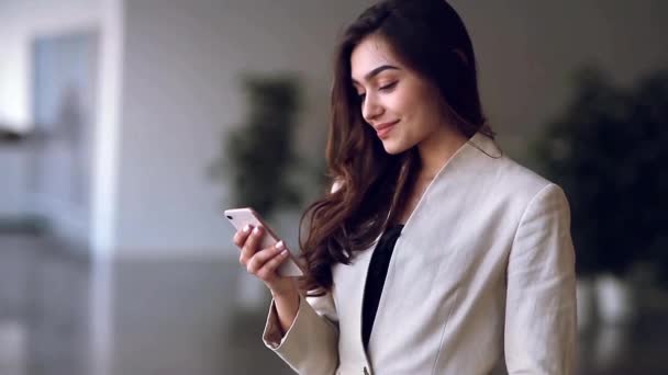 Beautiful Russian business girl with a phone in her hands, smiling. Office style, strict clothing and expressive look. Looks at the phone and laughs - Video