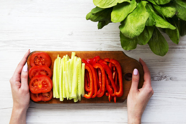 A girl is holding a board with freshly sliced vegetables: tomato, cucumber and pepper. Washed fresh bunch of spinach leaves . Top view. - Photo, Image