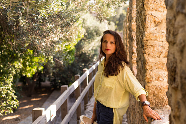 Self-confident brunette female wearing cotton yellow shirt, large watch and dark blue trousers standing by medieval castle wall against blurred olive trees. - Photo, Image