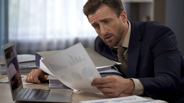 Exhausted man surrounded by paperwork at office looking at graphs, deadline - Photo, Image