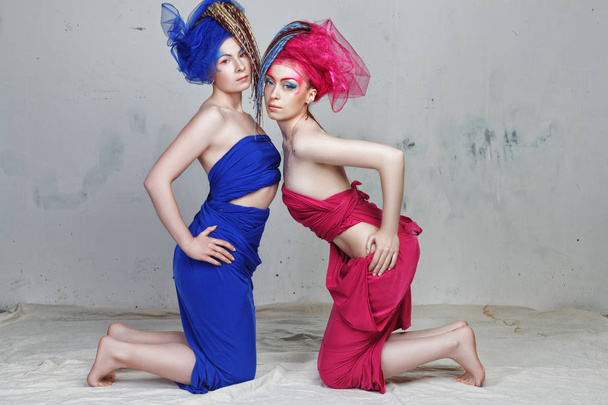 portrait of two girlfriends: girls with dreadlocks in grotesque dresses of blue and red are on their knees - Photo, Image