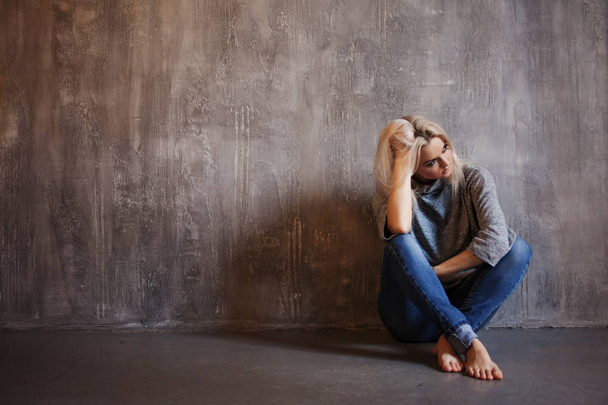 Sad woman sits on the floor. Depression and chronic fatigue. Young beautiful blonde in a gray sweater and jeans - Foto, Imagen