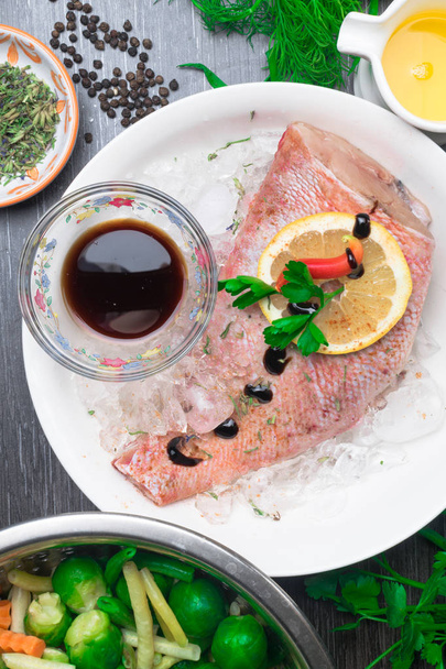 Whole raw organic fish sea perch on ice cubes in white plate on a dark table background with black pepper, olive oil, dill, cilantro, hyssop spices, parsley, soy sauce, lemon and decorated with red sweet pepper. Metal bowl with salad of green beans,  - Фото, изображение