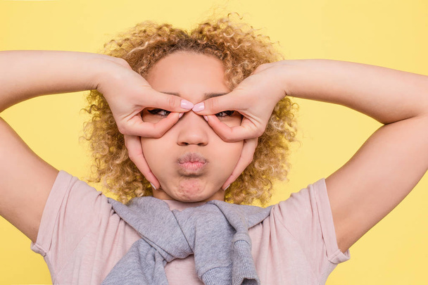 A picture of funny girl putting her fingers in a circle position and holding some air in her mouth. She looks funny but serious. Isolated on yellow background. - 写真・画像