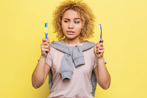 Girl is holding two teeth brushes in her hands and looking at one of them. Her sight is suspicious. She doesnt know what teeth brush should she use. Isolated on yellow background - Photo, Image
