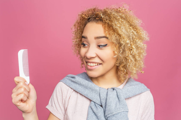 Pretty girl is looking to the brush that she holds in her hands and smiling. She cares about her blonde and curly hair. Isolated on pink background. - Photo, Image