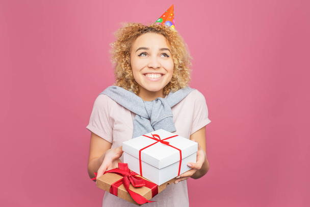 Happy girl is holding some presents in her hands. Also she has a birthday hat in her head. She is looking up and smiling. Young woman looks adorable. Isolated on pink background. - Foto, Imagen