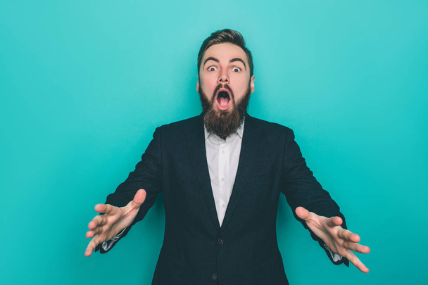 Berded man is absolutely amazed. He is looking to camera awith his mouth opened. Also he is showing his hands. Isolated on blue background. - 写真・画像