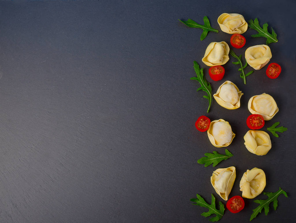 Frozen tortellini on the black background. Italian tortellini with fresh ricotta leaves and tomatoes on a black stone board. Top view. - Photo, Image