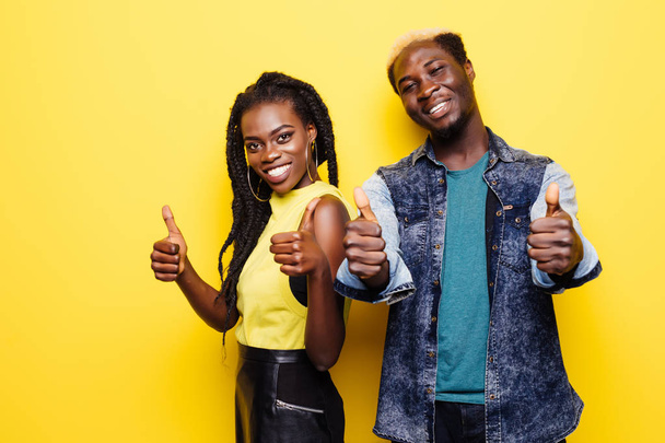 Portret van een lachende jonge afro Amerikaanse echtpaar permanent en tonen duimschroef opwaarts gebaar geïsoleerd op gele achtergrond - Foto, afbeelding