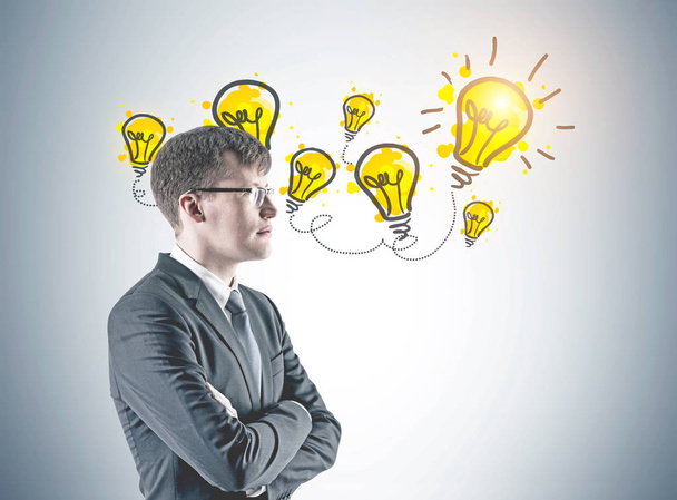Side view of a confident young businessman wearing a suit and glasses and standing with crossed arms looking into the distance. Yellow light bulbs on a gray wall. An idea concept - Zdjęcie, obraz