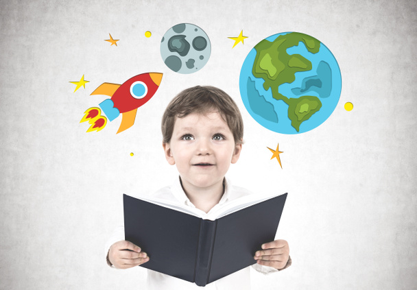 Netter kleiner Junge in weißem Hemd, der ein Buch in der Nähe einer Betonwand hält, auf dem helle Raketen-, Erd- und Mondzeichnungen stehen.. - Foto, Bild