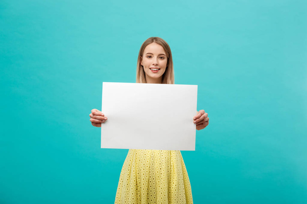 Lifestyle Concept: young beautiful girl smiling and holding a blank sheet of paper, dressed in yellow, isolated on pastel blue background - Photo, Image