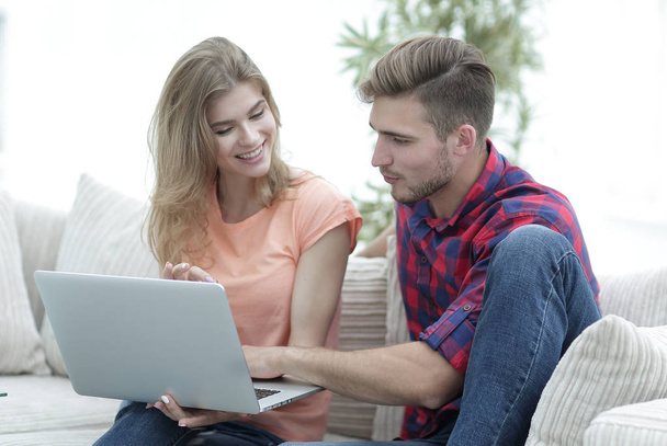 若いカップルはラップトップを使用して、自宅のソファに腰掛けながら笑顔 - 写真・画像