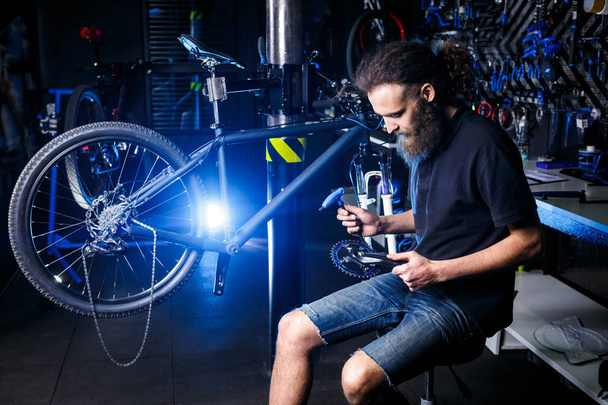 Молодой стильный бородатый, с длинными волосами кавказский механик-мужчина в велосипедной мастерской использует инструмент для ремонта комплектов Cranksets
 - Фото, изображение