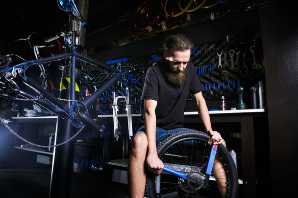 Giovane elegante barba, con i capelli lunghi maschio meccanico caucasico in un laboratorio di biciclette utilizza uno strumento per riparare cassette sulla ruota posteriore di una bicicletta
 - Foto, immagini