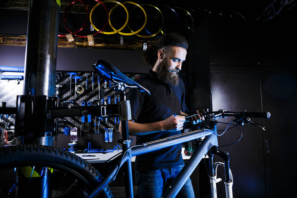 Jeune homme barbu élégant aux cheveux longs Homme caucasien mécanicien travailleur dans un atelier de vélo utilise un outil pour réparer un vélo Bars avec une clé hexagonale
 - Photo, image