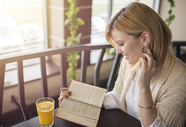 Joven mujer rubia sonriente en un restaurante leyendo un libro y dr
 - Foto, Imagen