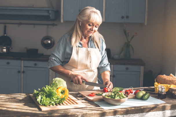 Serene mature lady cutting vegetables and enjoying song - Foto, afbeelding