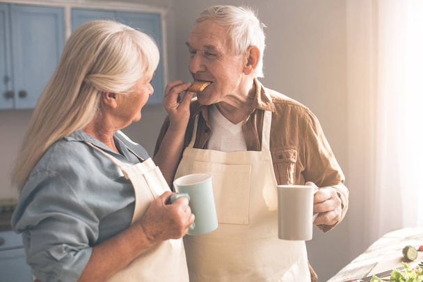Happy senior couple drinking hot beverage with sweets at home - Foto, Imagem