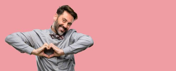 Hombre de mediana edad, con barba y pajarita feliz mostrando amor con las manos en forma de corazón expresando saludable y símbolo de matrimonio
 - Foto, Imagen