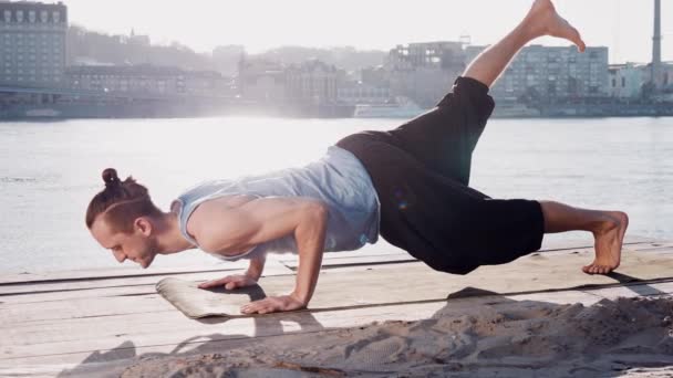 junger kaukasischer Mann entspannt sich durch Yoga-Fitness-Übungen am Strand in der Nähe des ruhigen Flusses mit Stadt im Hintergrund - Filmmaterial, Video