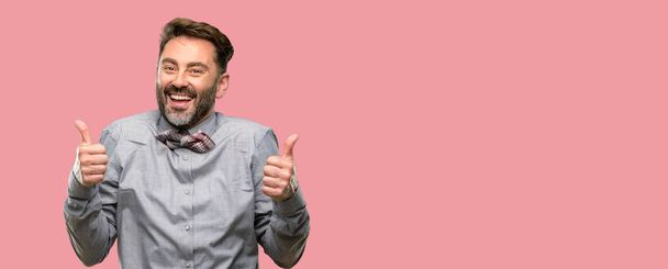 Мужчина средних лет, с бородой и галстуком-бабочкой широко улыбается показывая большие пальцы вверх жест к камере, выражение, как и и одобрение
 - Фото, изображение